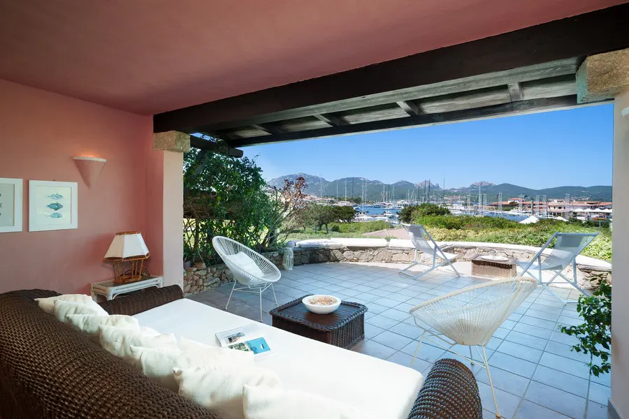 Outdoor patio with brown wicker couch, white cushions, and white chairs overlooking a harbor with boats and mountains in the distance.