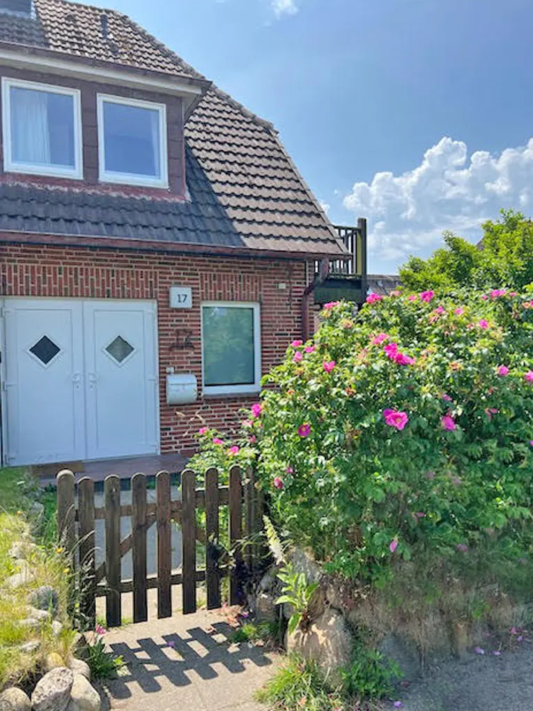 A red brick house with white doors and a brown tiled roof, number 17, is surrounded by green bushes with pink flowers.