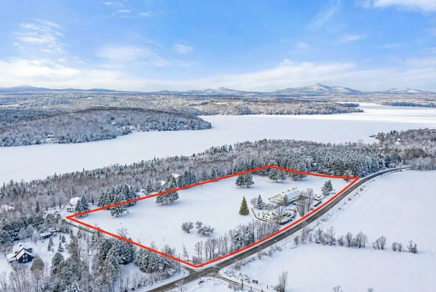 Aerial view of a snow-covered property outlined in red, with a house, trees, and a frozen lake in the background.