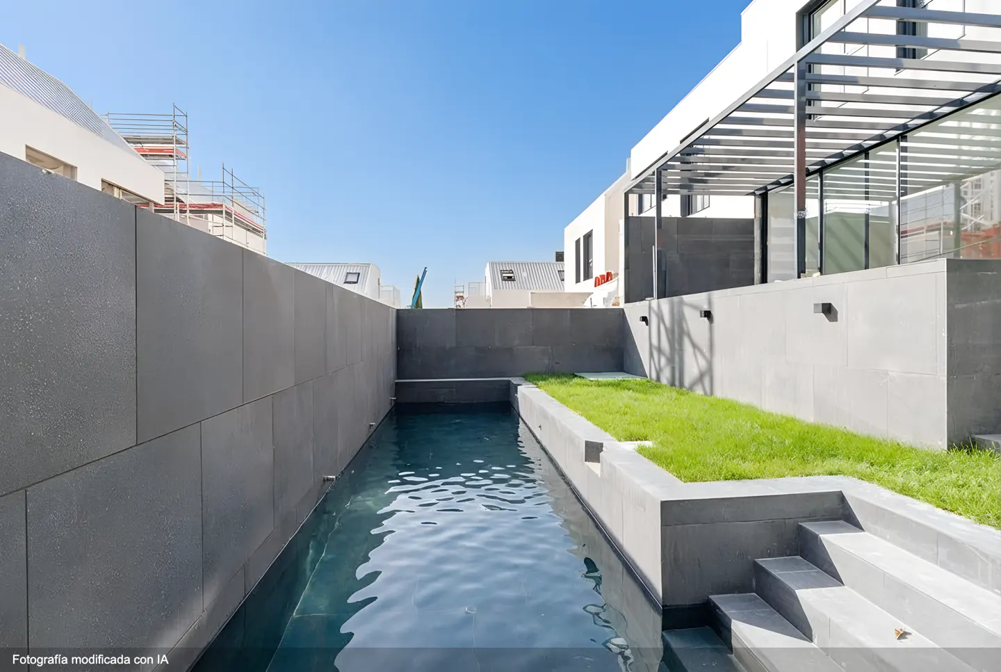 A modern backyard with a narrow pool, gray stone walls, and a small patch of green grass. The sky is blue and clear.
