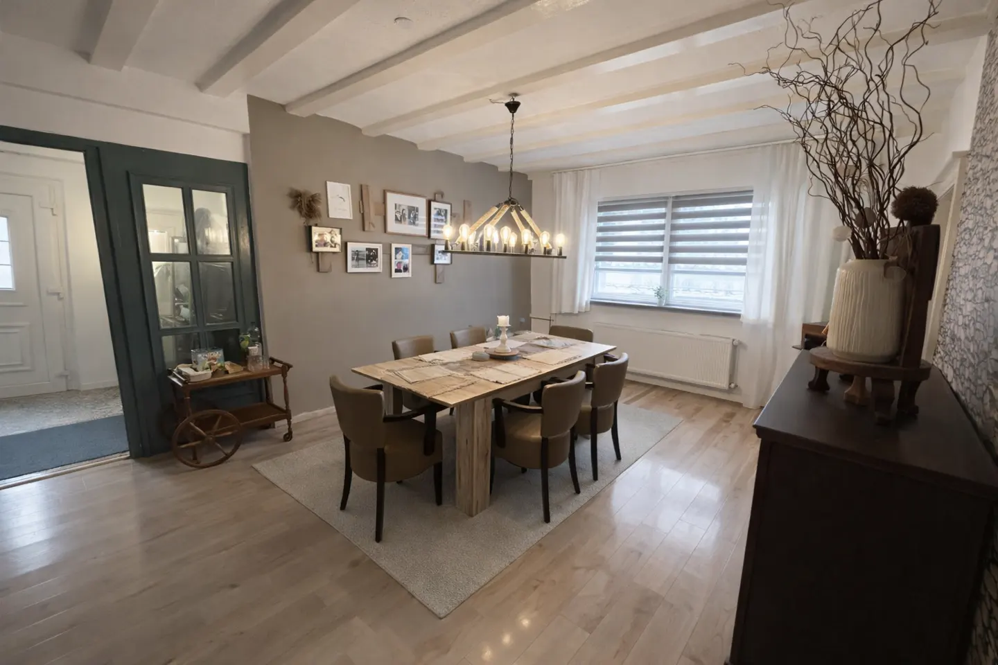 A dining room with a long wooden table, brown chairs, and a decorative light fixture. A gray wall with framed photos is visible.