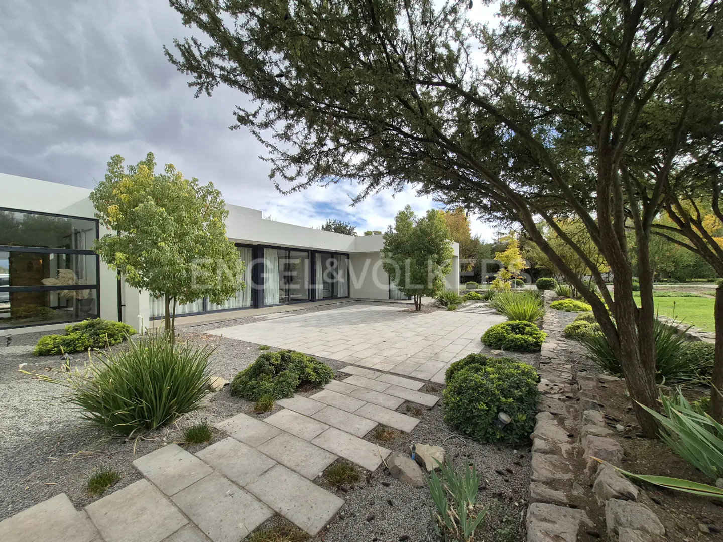 Modern white house with a stone patio, green trees, and bushes. The sky is cloudy. Engel & Volkers logo is visible.