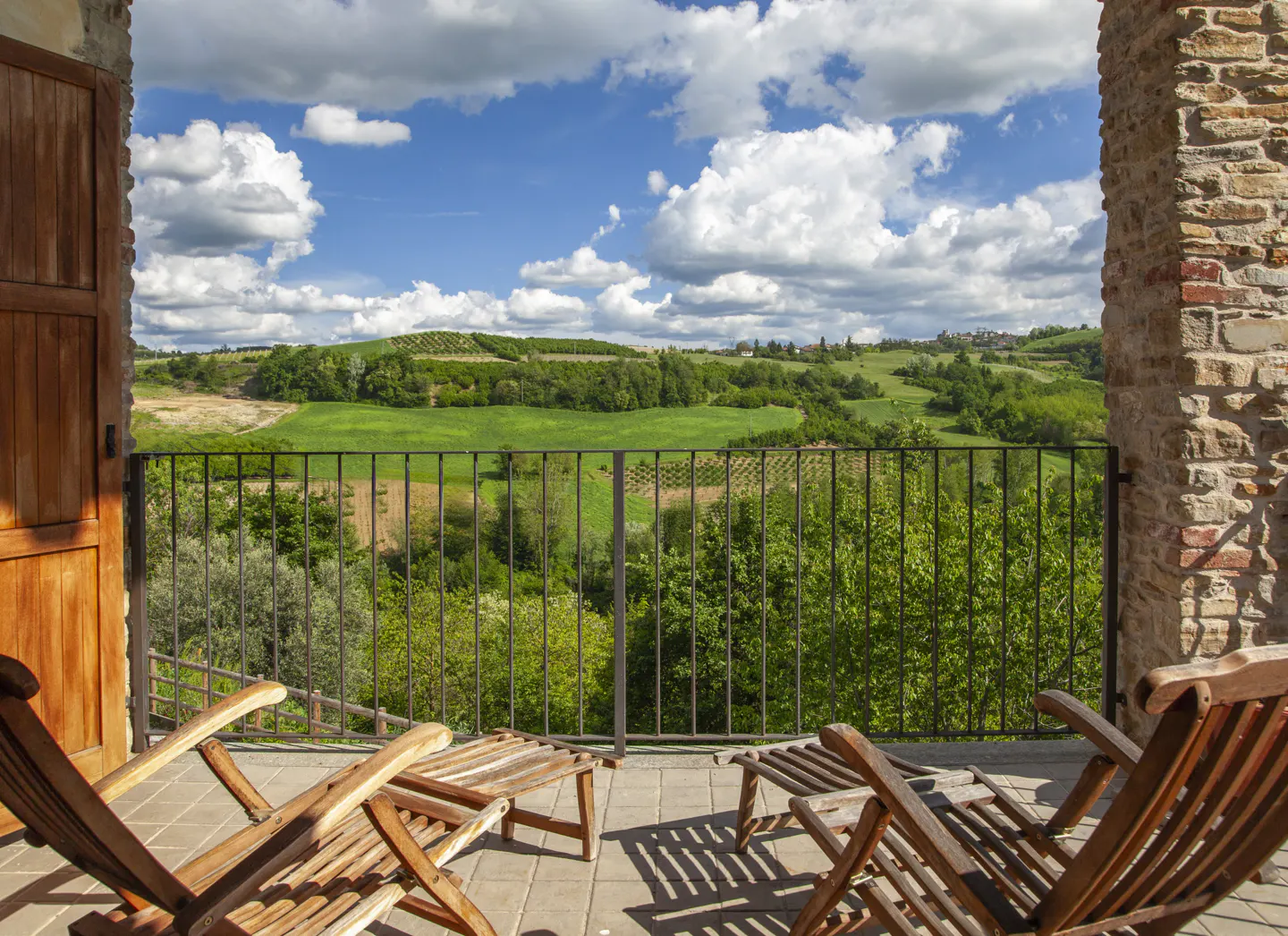 Balcony view with two wooden lounge chairs overlooking green hills and a blue sky with white clouds.