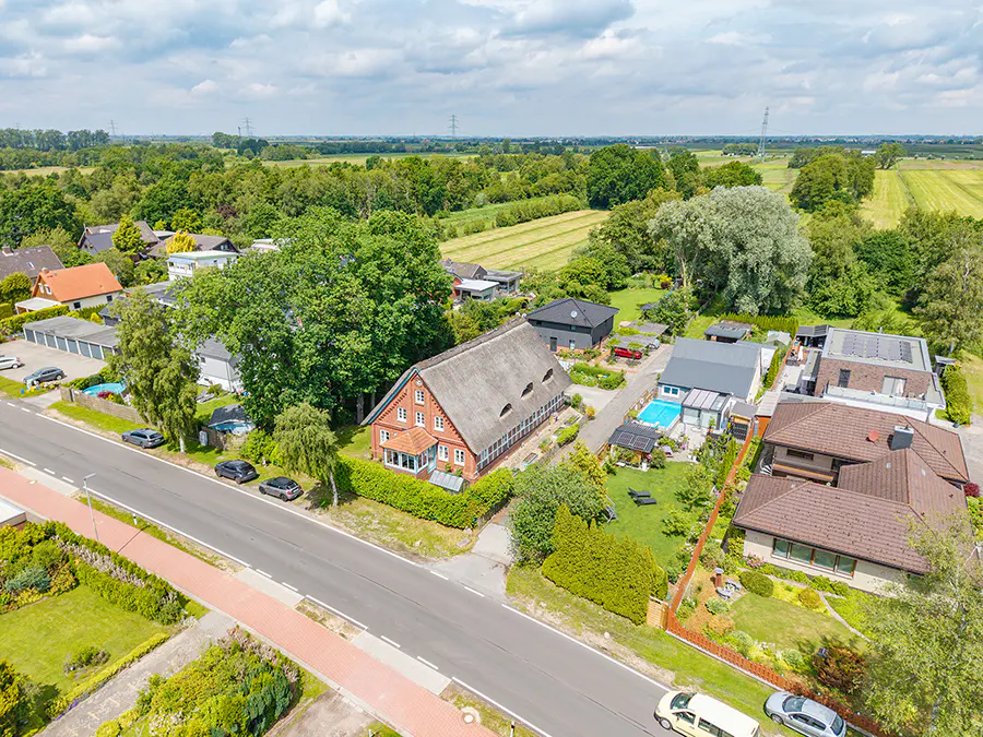 Aerial view of a red brick house with a thatched roof in a lush, green residential neighborhood with nearby swimming pools.