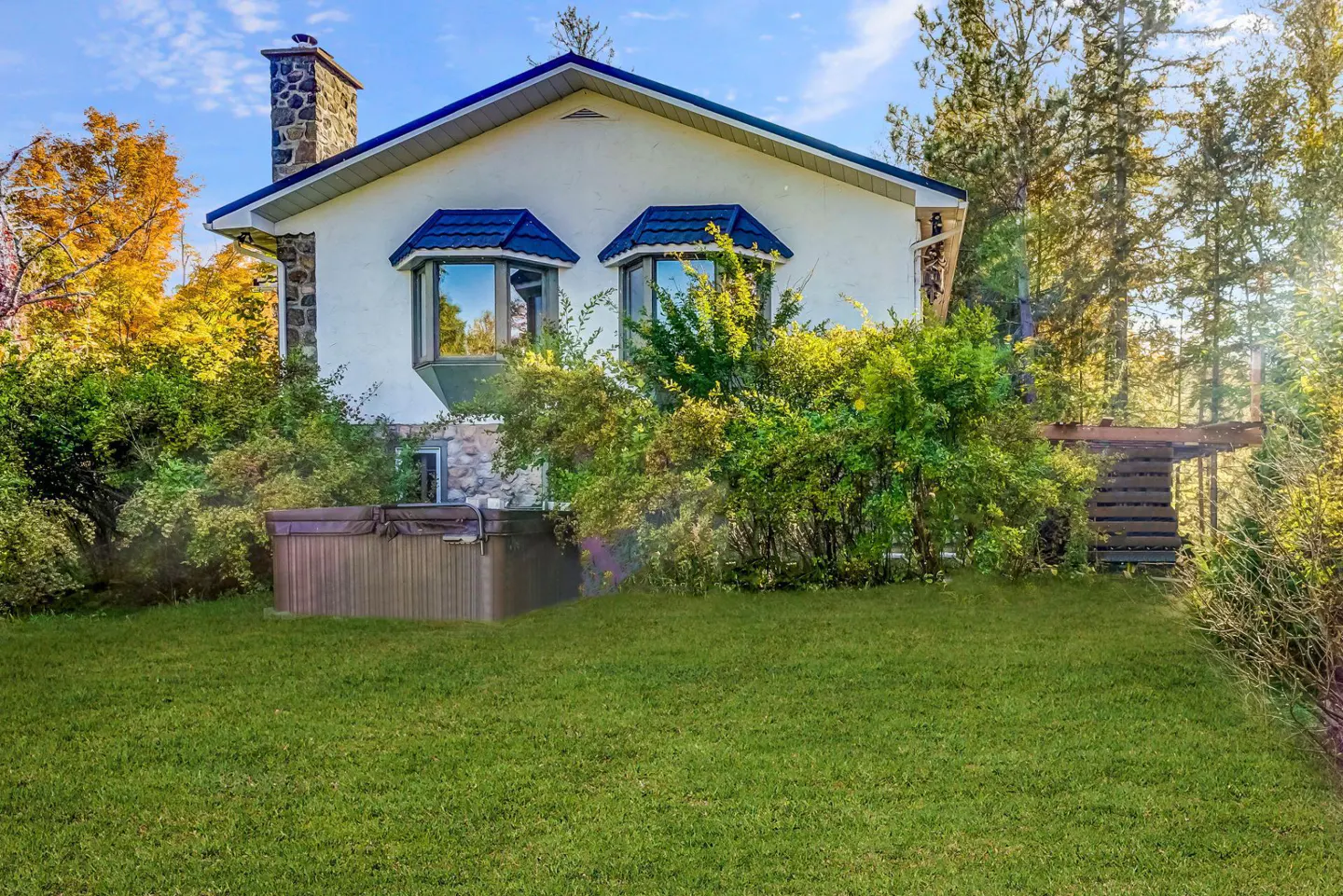 A white house with blue trim and a stone chimney, surrounded by green grass and trees.