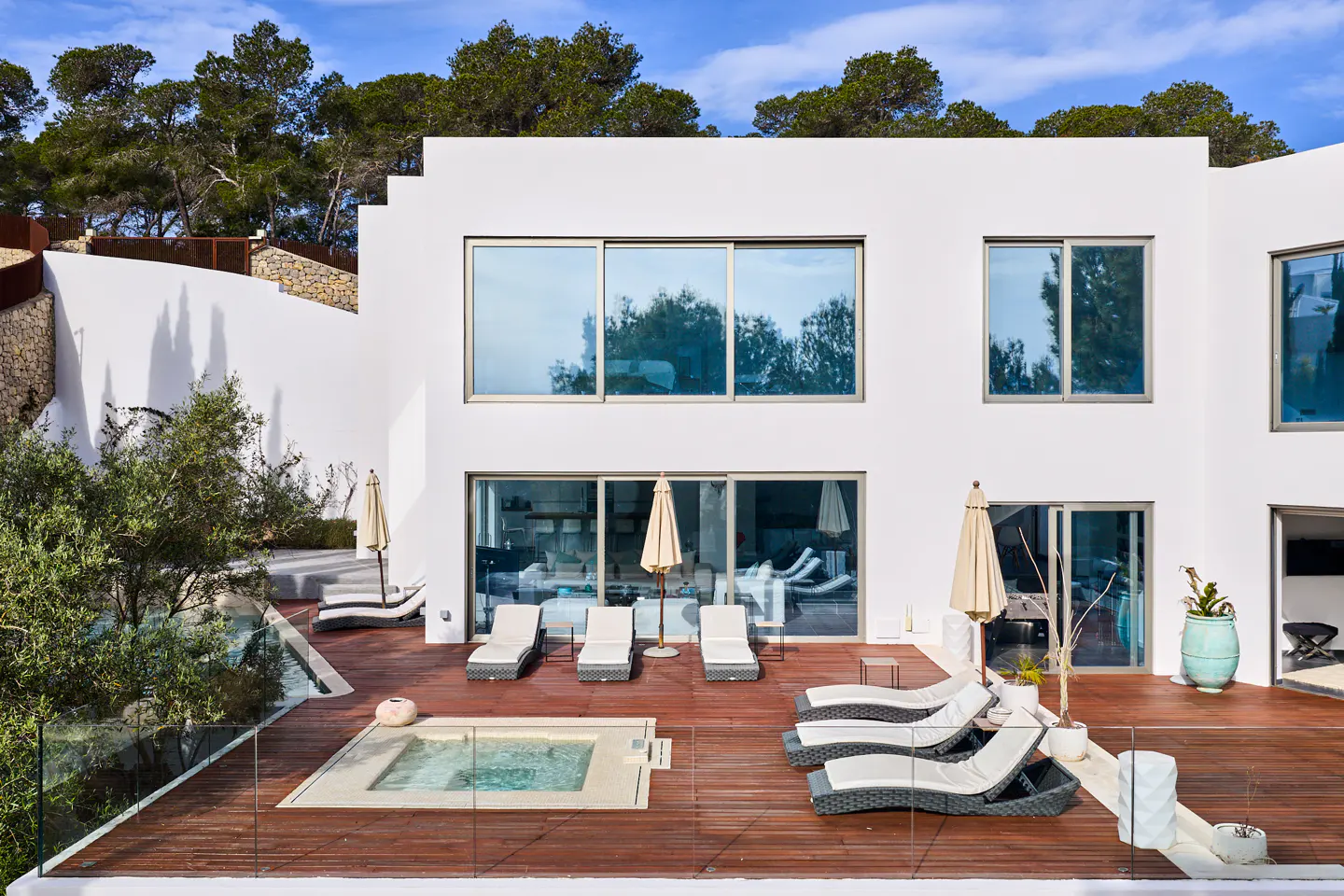 Modern white house with a wooden deck, lounge chairs, and a small pool. Trees and blue sky in the background.
