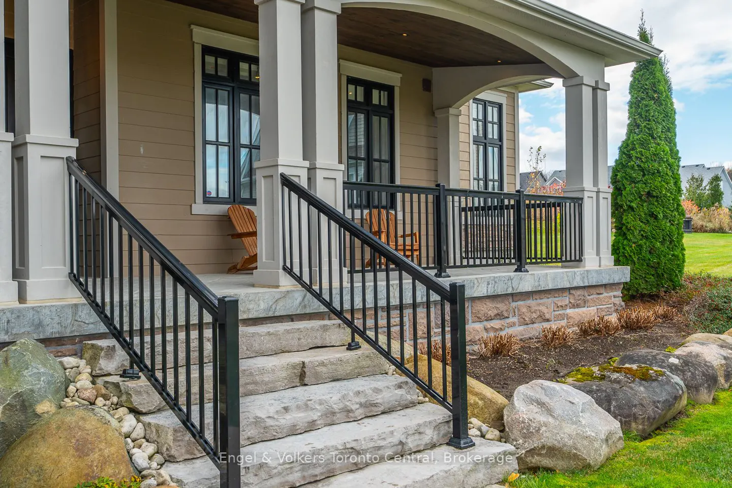 Exterior view of a house with stone steps, black railings, and a covered porch with wooden chairs. The house has beige siding and white columns.