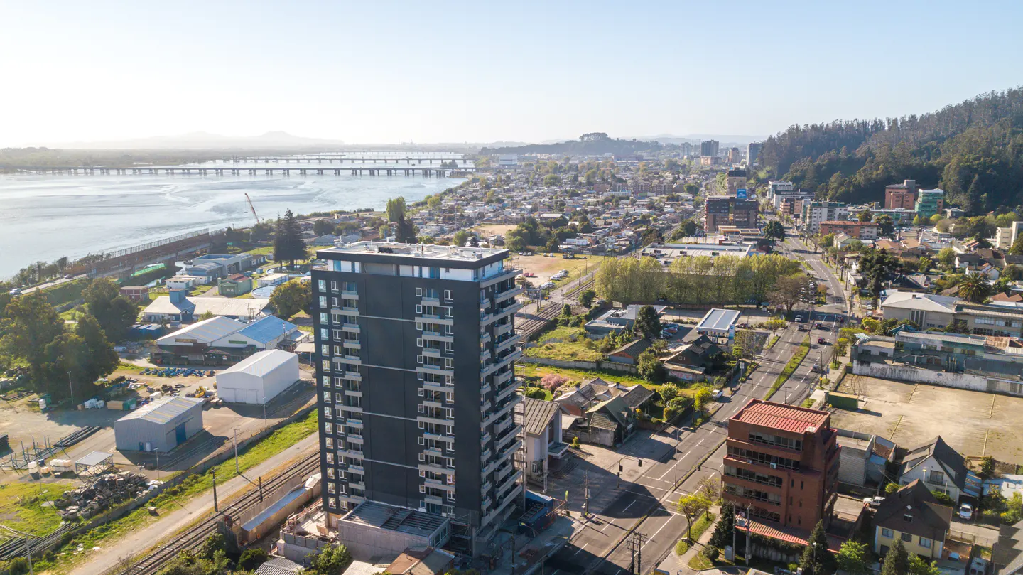 Aerial view of a city with a tall, dark gray apartment building in the foreground, a river with bridges, and mountains in the background.