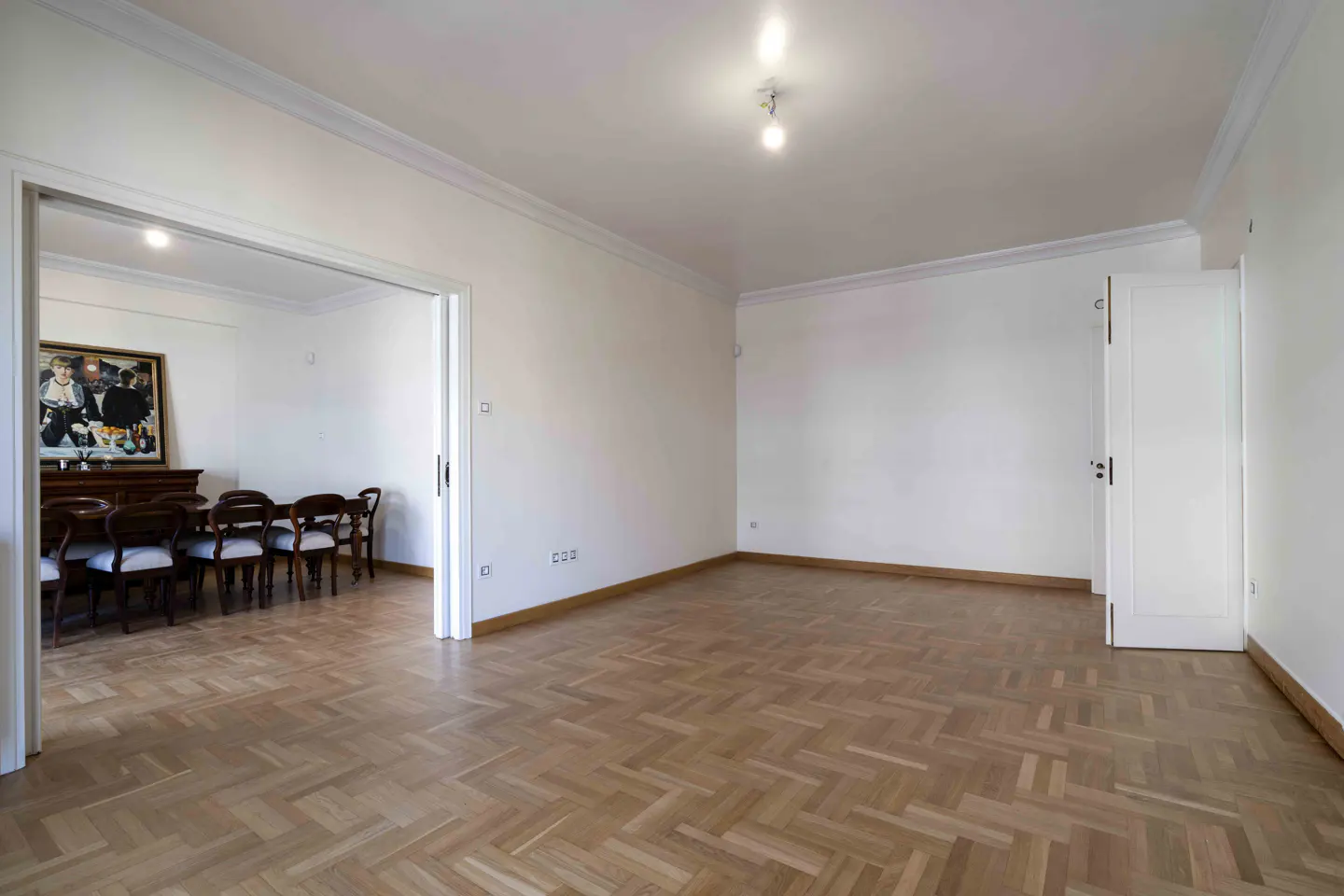Empty room with herringbone wood floors, white walls, and a doorway to a dining room with a table, chairs, and painting.