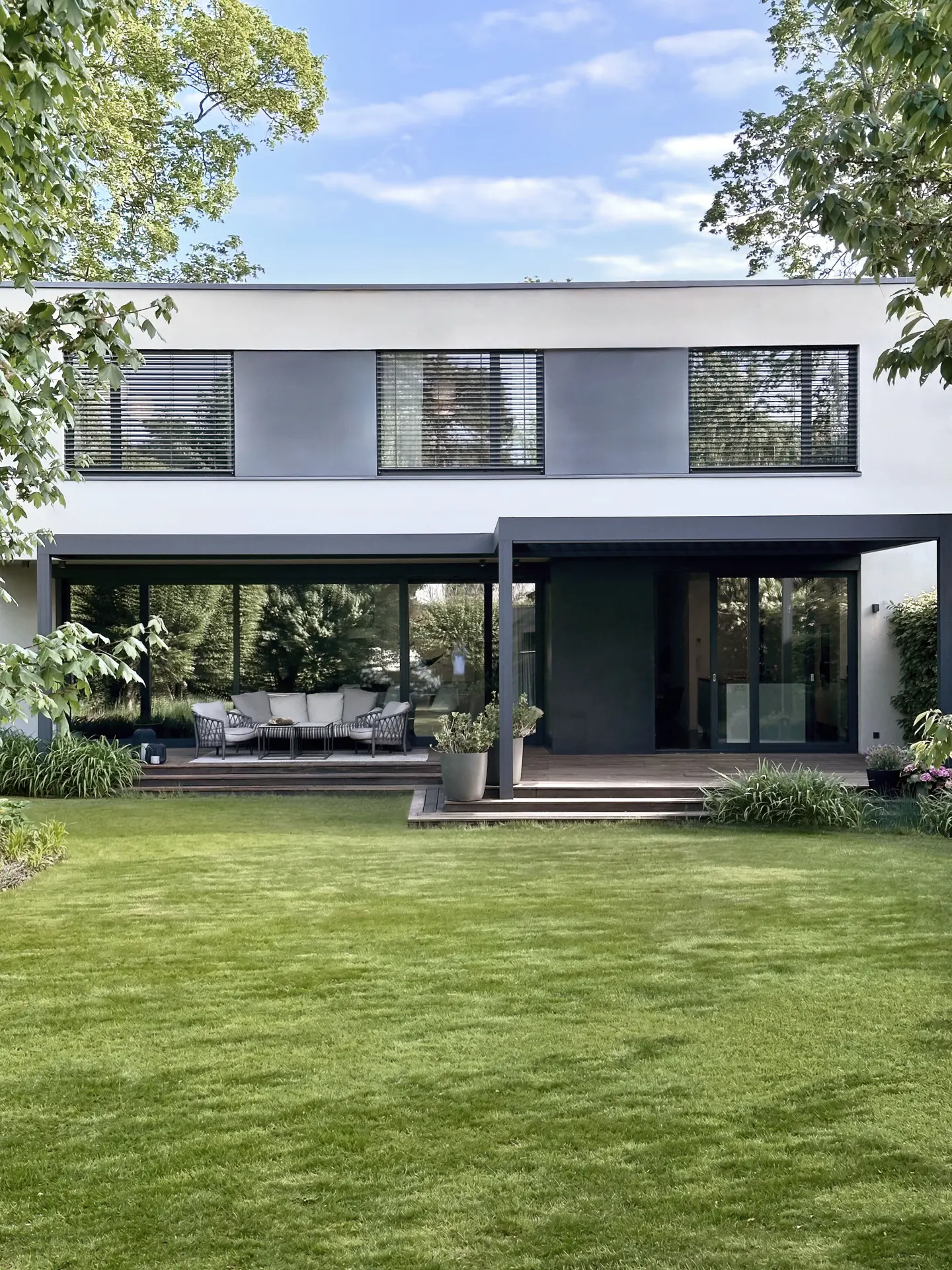 Modern two-story house with a large green lawn. The house has large windows and a patio with outdoor furniture.