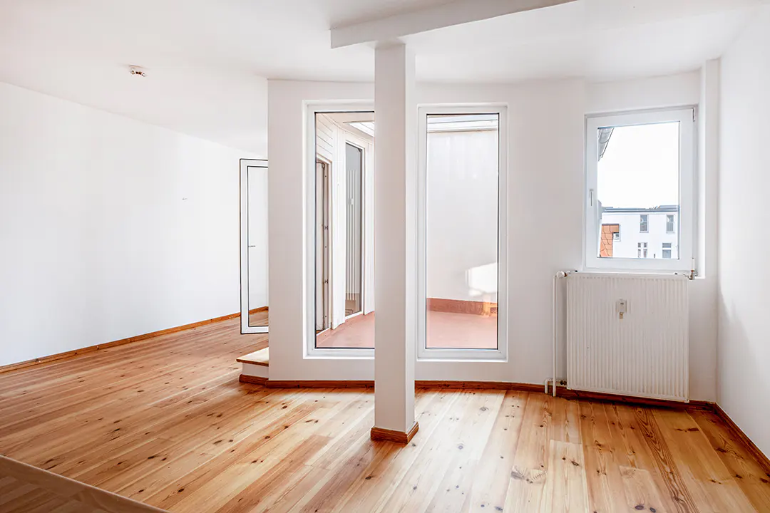 Bright, empty room with wood floors, white walls, and a white column. Windows look out to a balcony and a building.