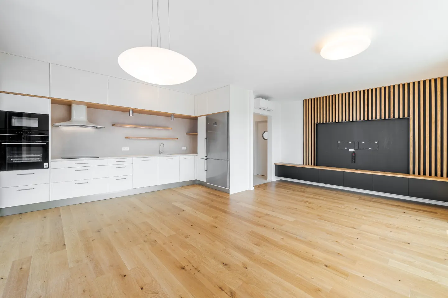 A modern apartment interior with white cabinets, stainless steel appliances, and a wood-slatted wall with a large TV.