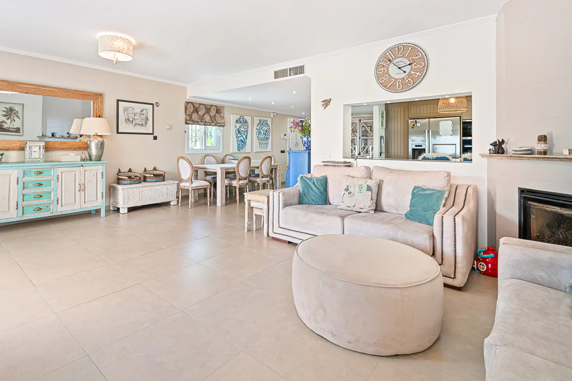 A bright living room with a beige sofa, round ottoman, fireplace, and dining area in the background.