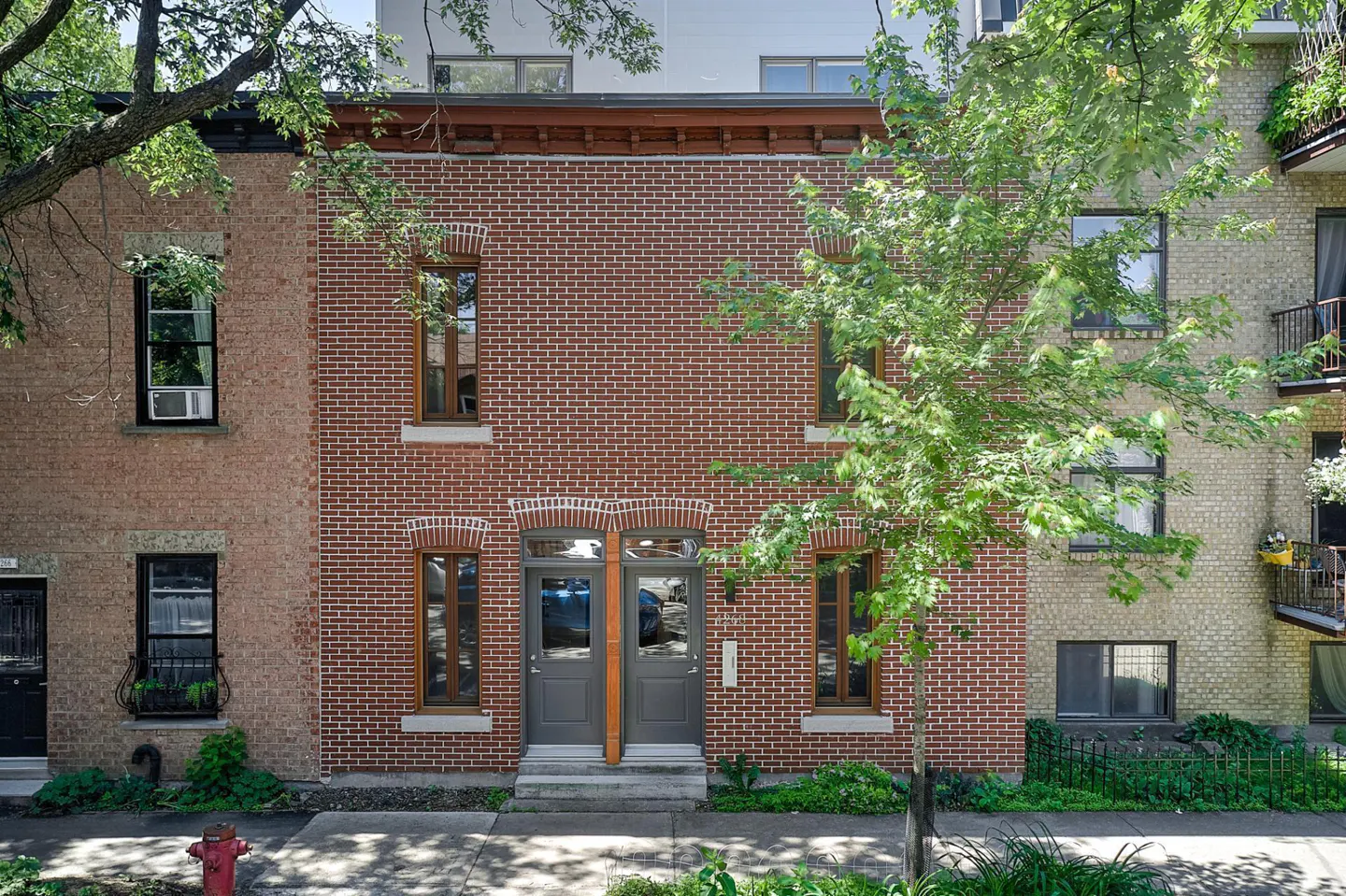 Red brick townhouse with two grey front doors, framed windows, and a tree in front on a sunny city street.