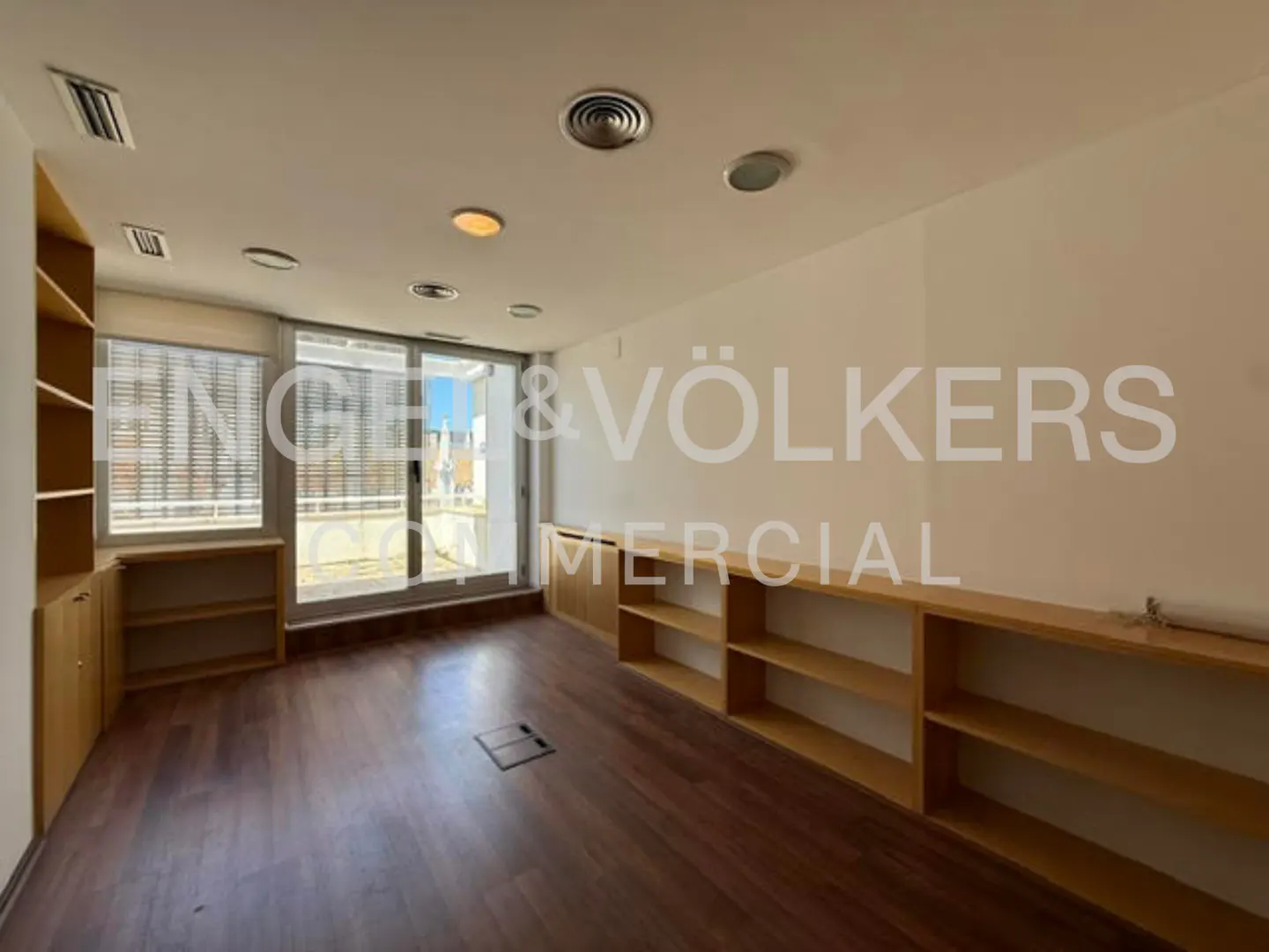 Empty office with wooden floors, built-in light wood shelving, and large glass doors leading to a bright balcony.