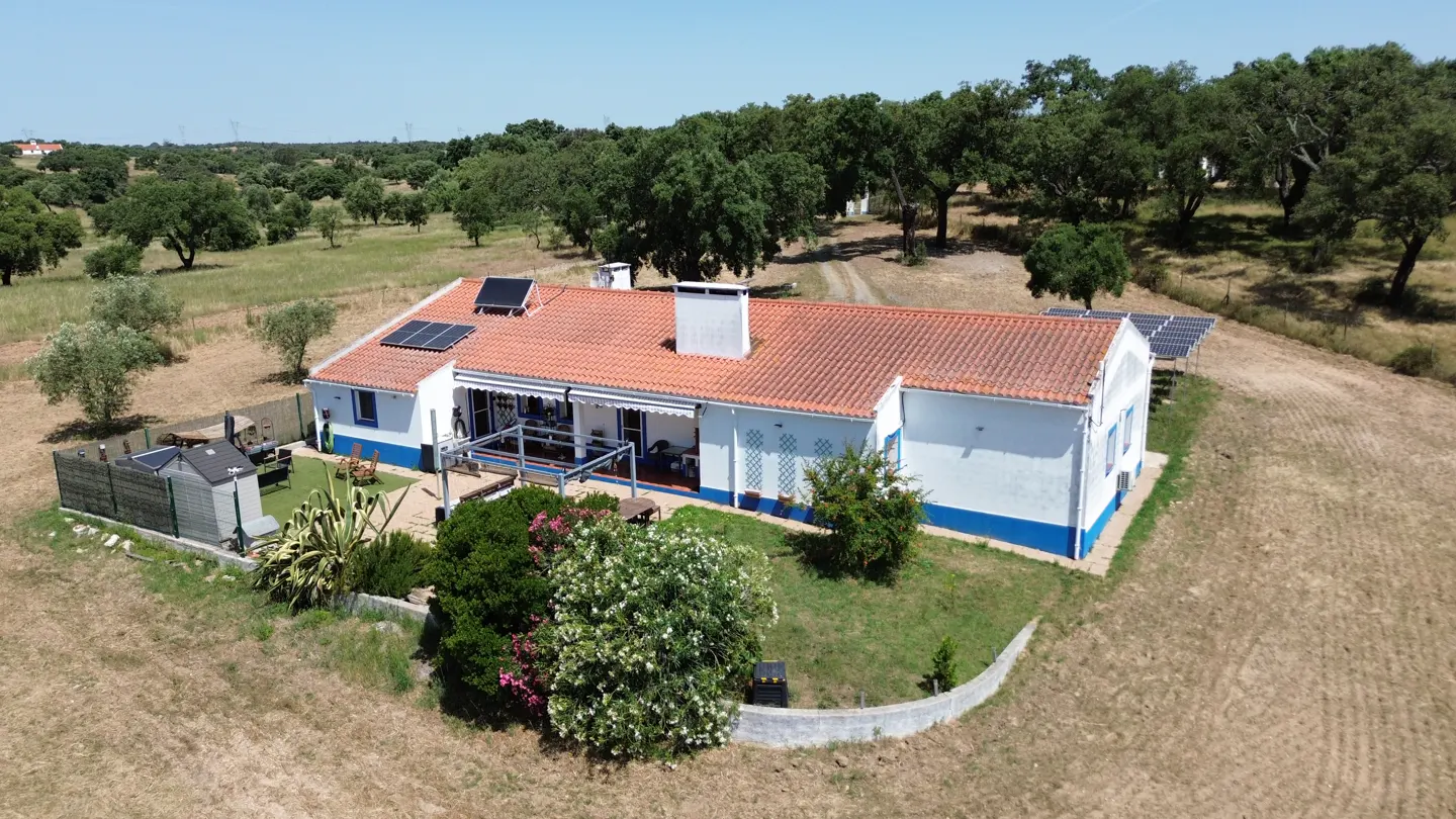 Aerial view of a white house with a red tile roof, solar panels, and a patio, set in a rural landscape with oak trees.