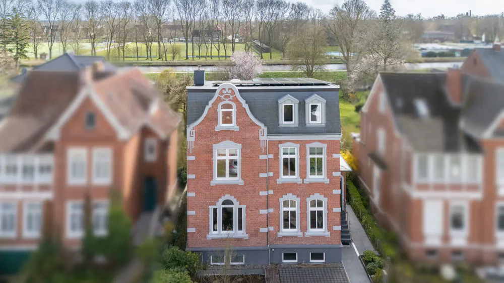 A three-story red brick house with white trim and a gray roof stands between two blurred houses. Solar panels are on the roof.