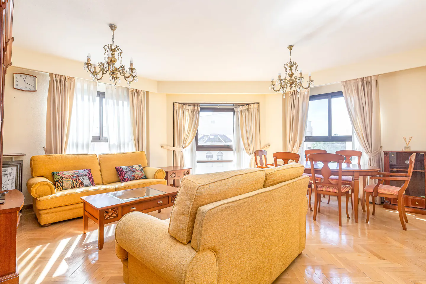 Bright living room with yellow sofas, wood floors, and dining table. Chandeliers hang from the ceiling. Windows offer a city view.