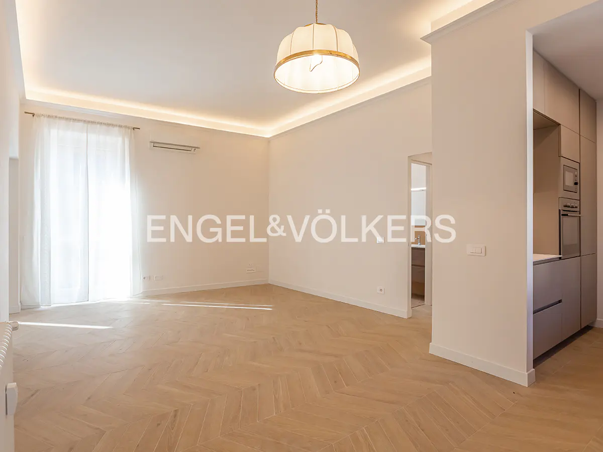 Bright, empty room with herringbone wood floors, white walls, and a modern light fixture. Kitchen cabinets are visible in the background.