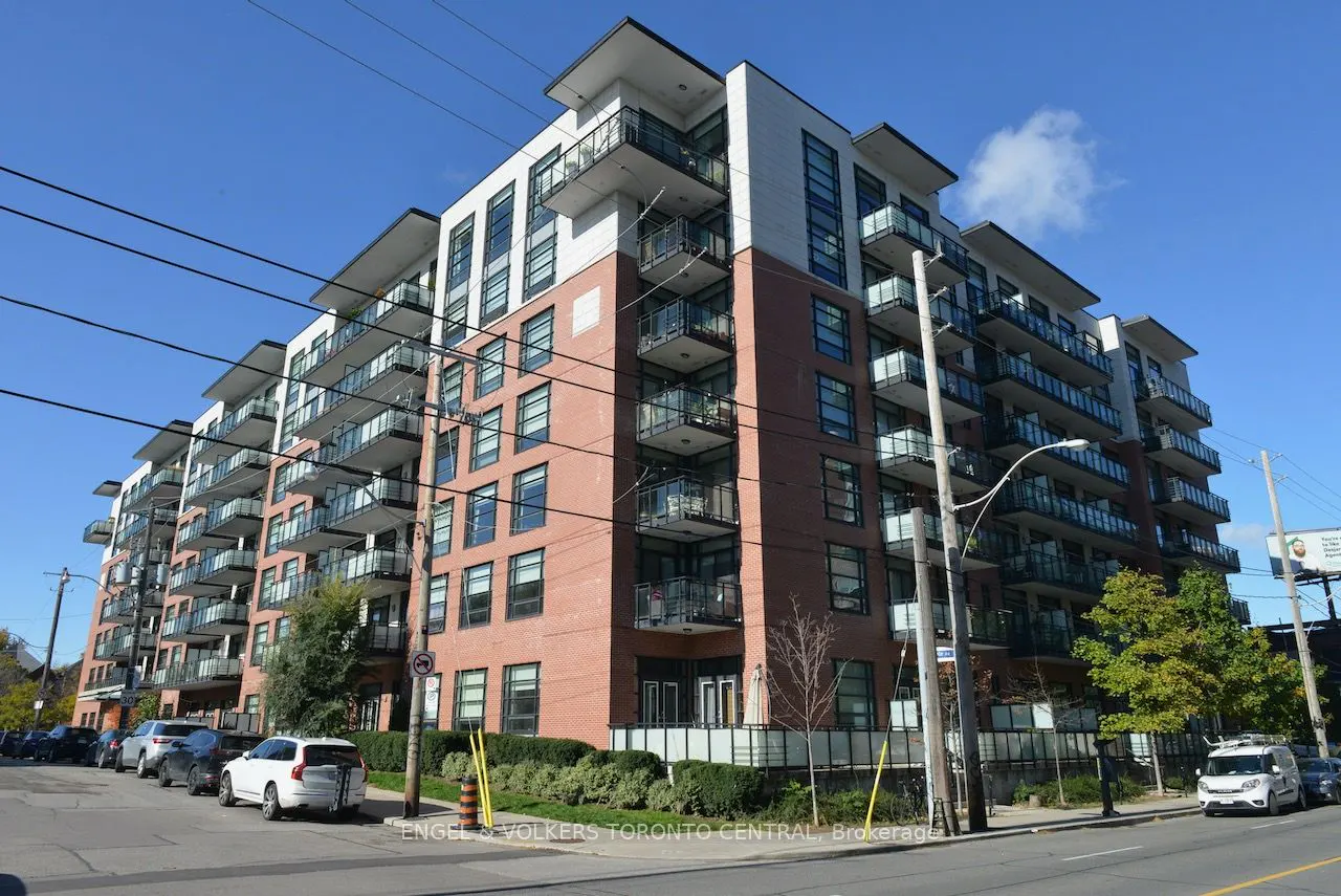Exterior view of a modern, multi-story brick apartment building with balconies on a sunny day. Cars are parked along the street.
