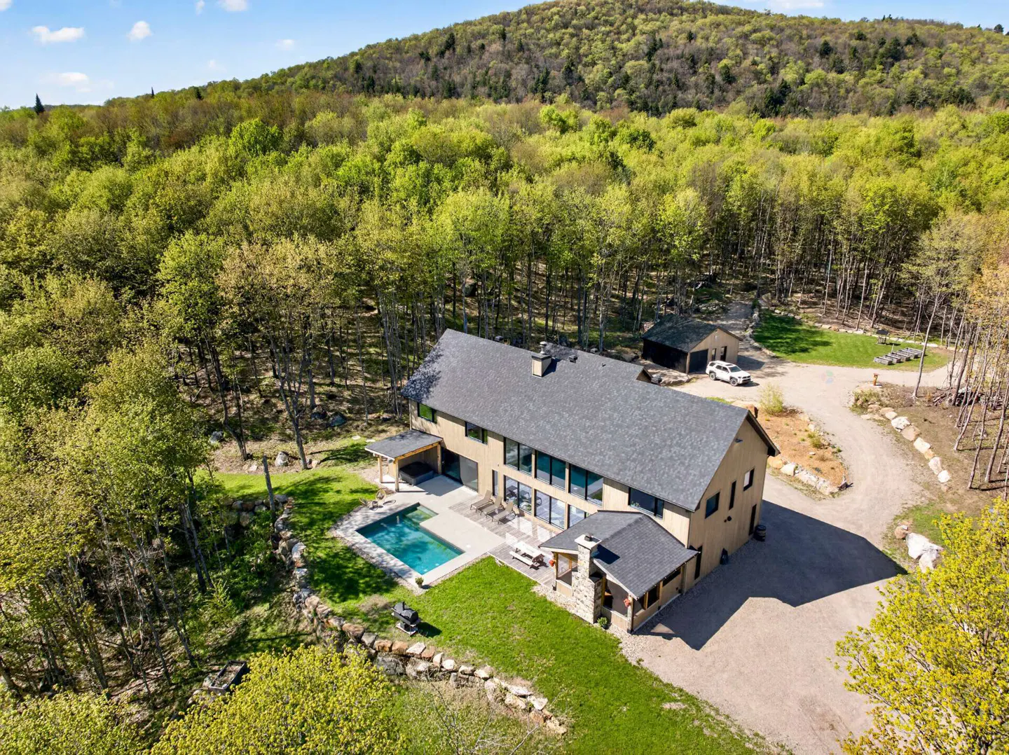Aerial view of a modern tan house with a pool, surrounded by green trees and a mountain backdrop.