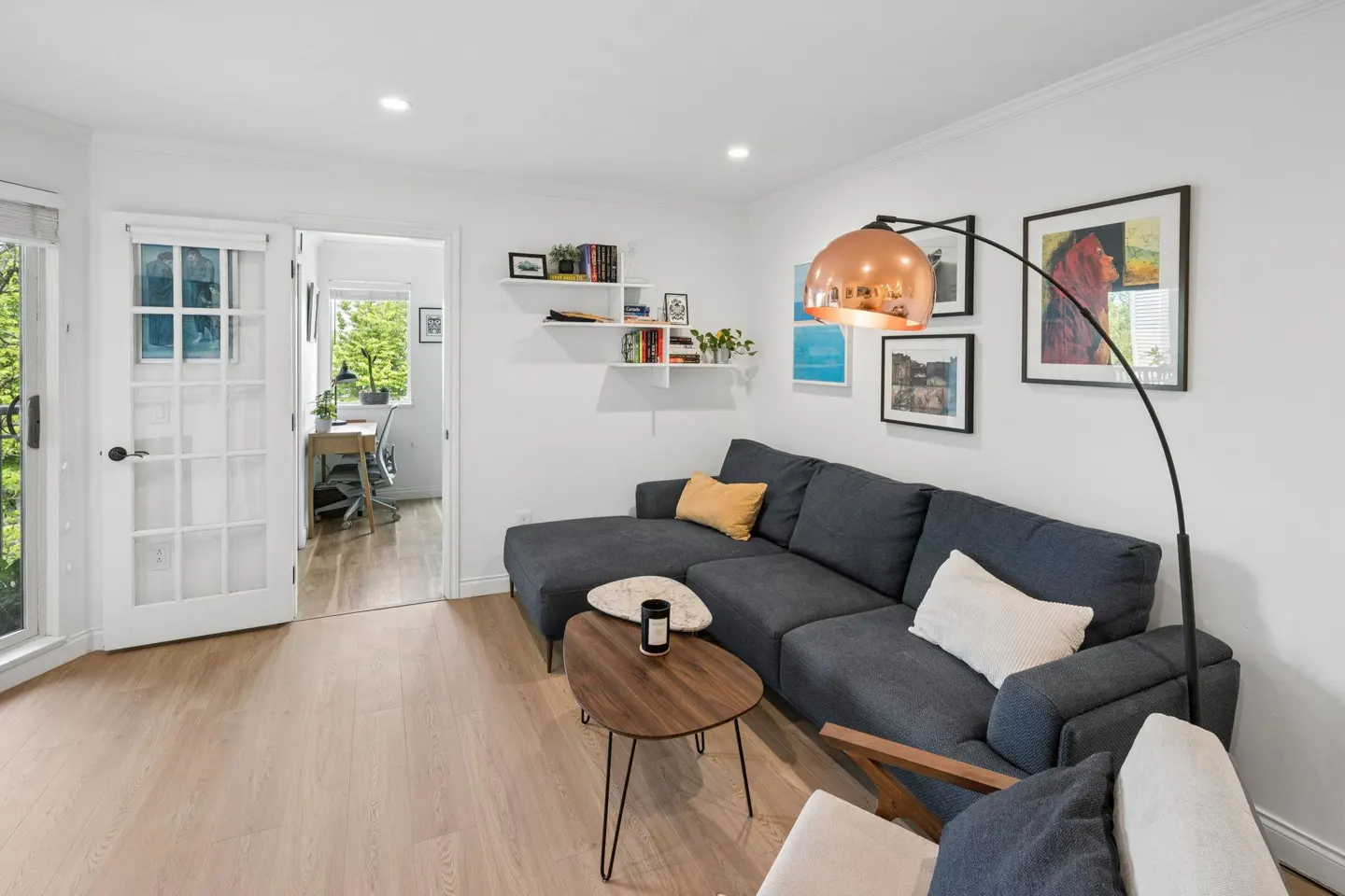 Bright living room with a gray sectional sofa, wood floors, and white walls. A copper floor lamp arches over the sofa. A doorway leads to an office.
