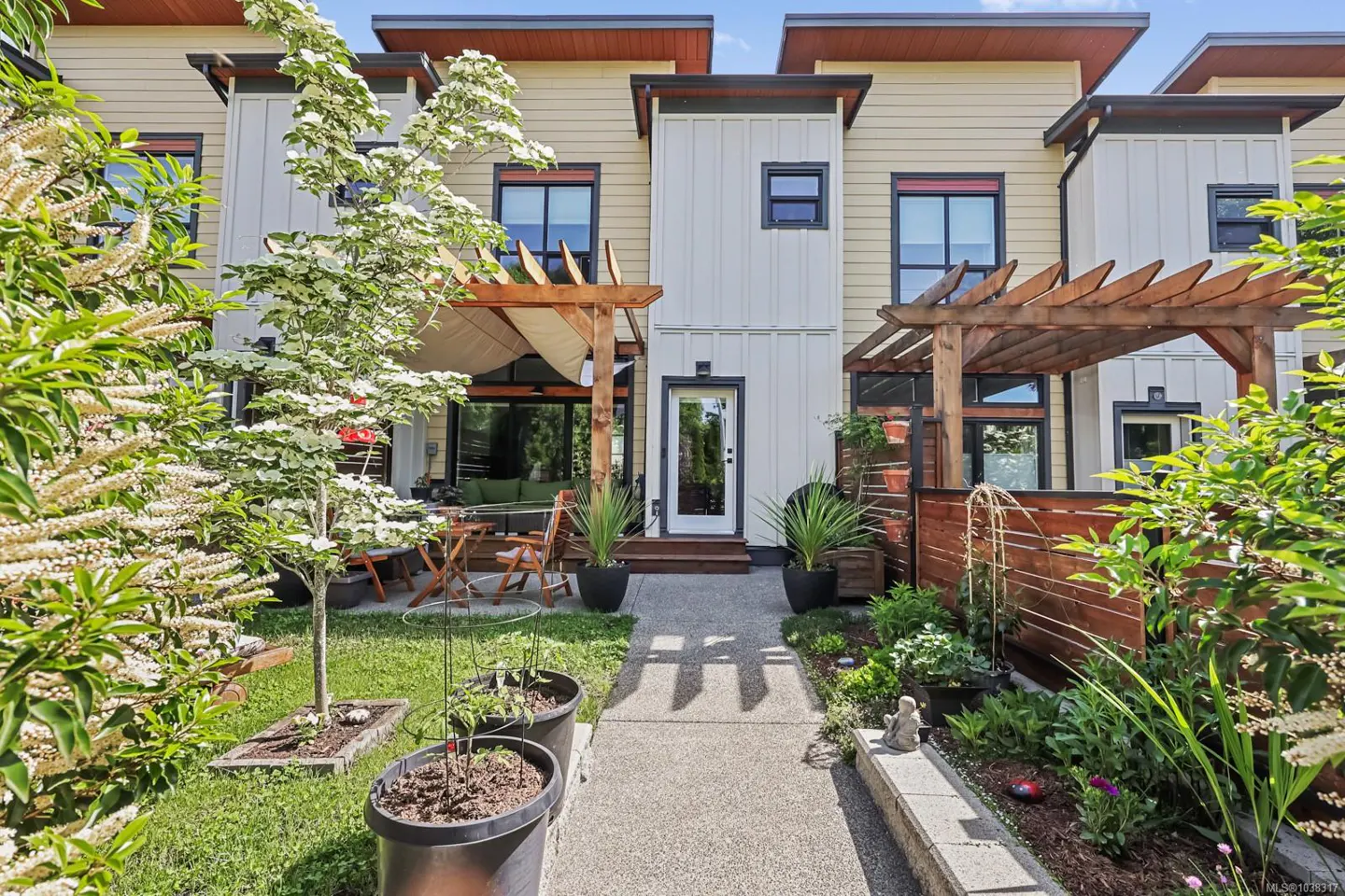 A concrete path leads to a modern townhouse patio with wooden pergolas, potted plants, and a small garden area.