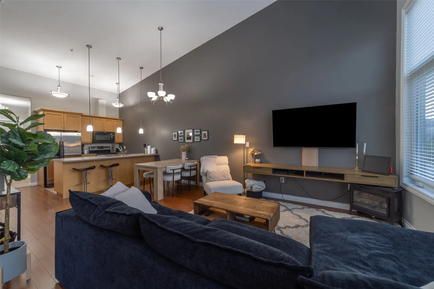Open-concept living space with a navy sofa, wood floors, and a gray accent wall. Kitchen with stainless steel appliances and a dining area.