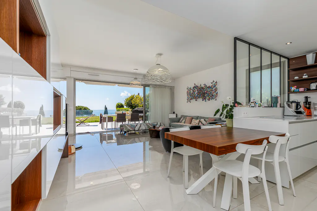 Bright, modern living space with white tile floors, a wood table with white chairs, and a view of the ocean through sliding glass doors.