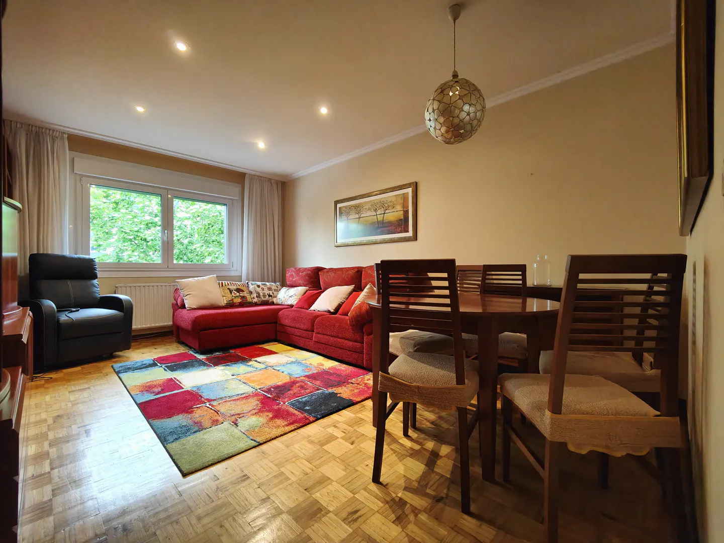A living room with a red sectional sofa, a colorful rug, and a wooden dining table with chairs. A black recliner sits near a window with white curtains.