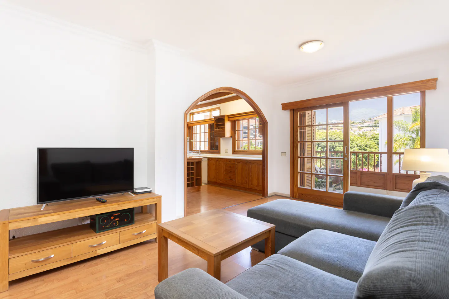 Living room with a grey sectional sofa, wooden coffee table, TV stand, and an arched doorway leading to a wooden kitchen.