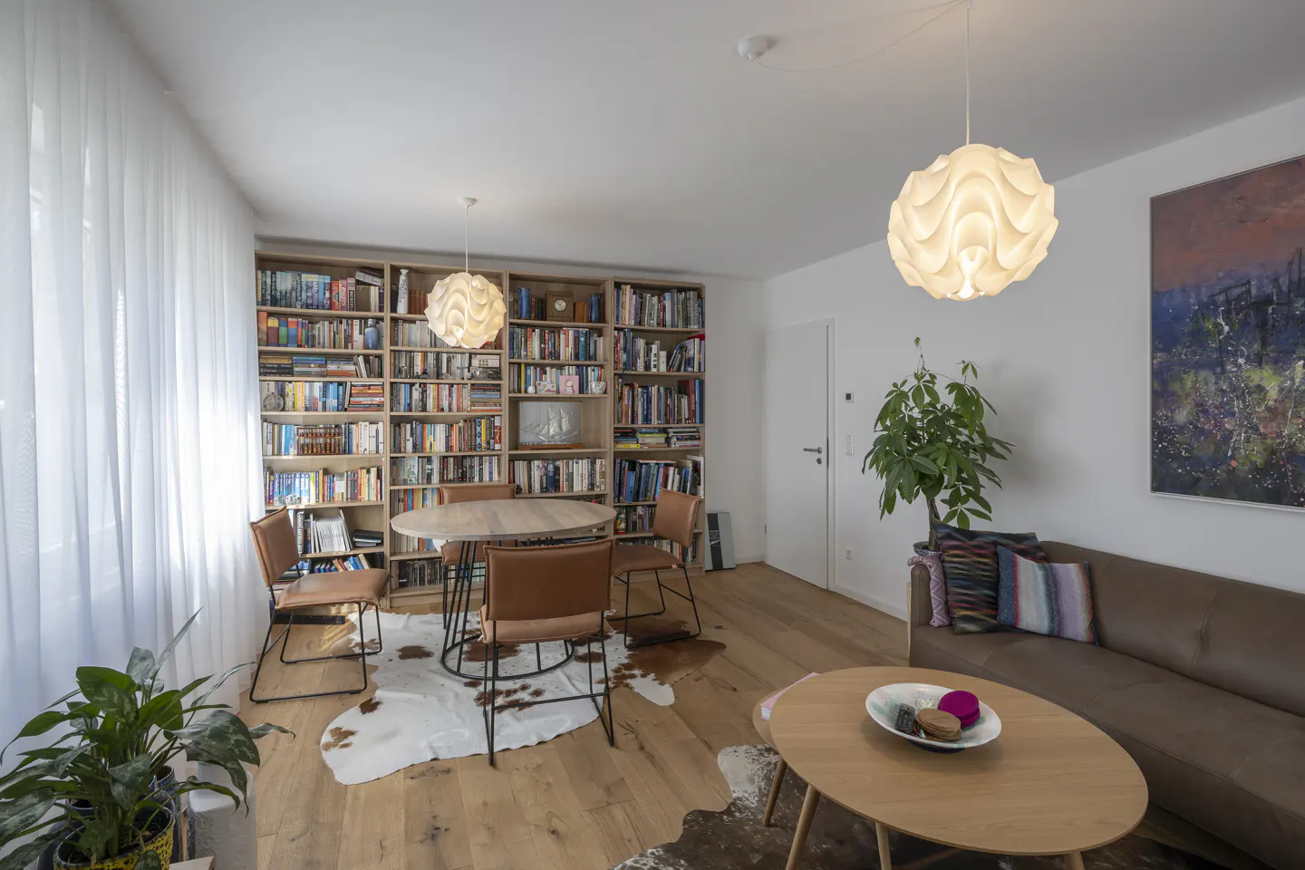 Bright living room with hardwood floors, a large bookshelf filled with books, a brown leather sofa, and a round wooden table with chairs.