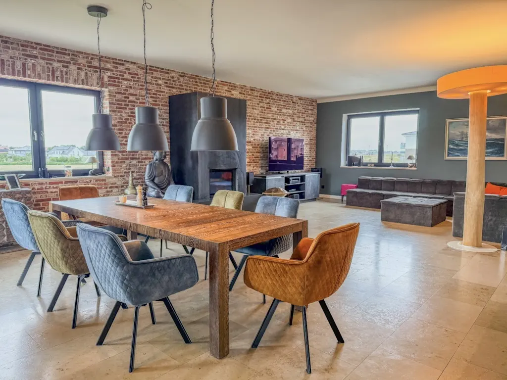 A modern living and dining area with a long wooden table, colorful chairs, and a brick wall.