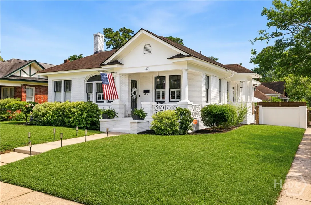 Charming white brick bungalow with a brown roof, front porch, American flag, and manicured lawn under a clear blue sky.