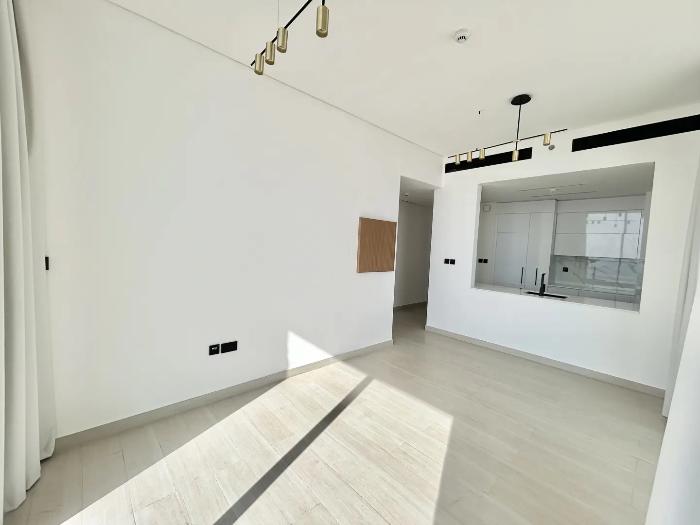 Bright, minimalist living room with white walls, light wood floors, modern gold pendant lights, and an open kitchen view.