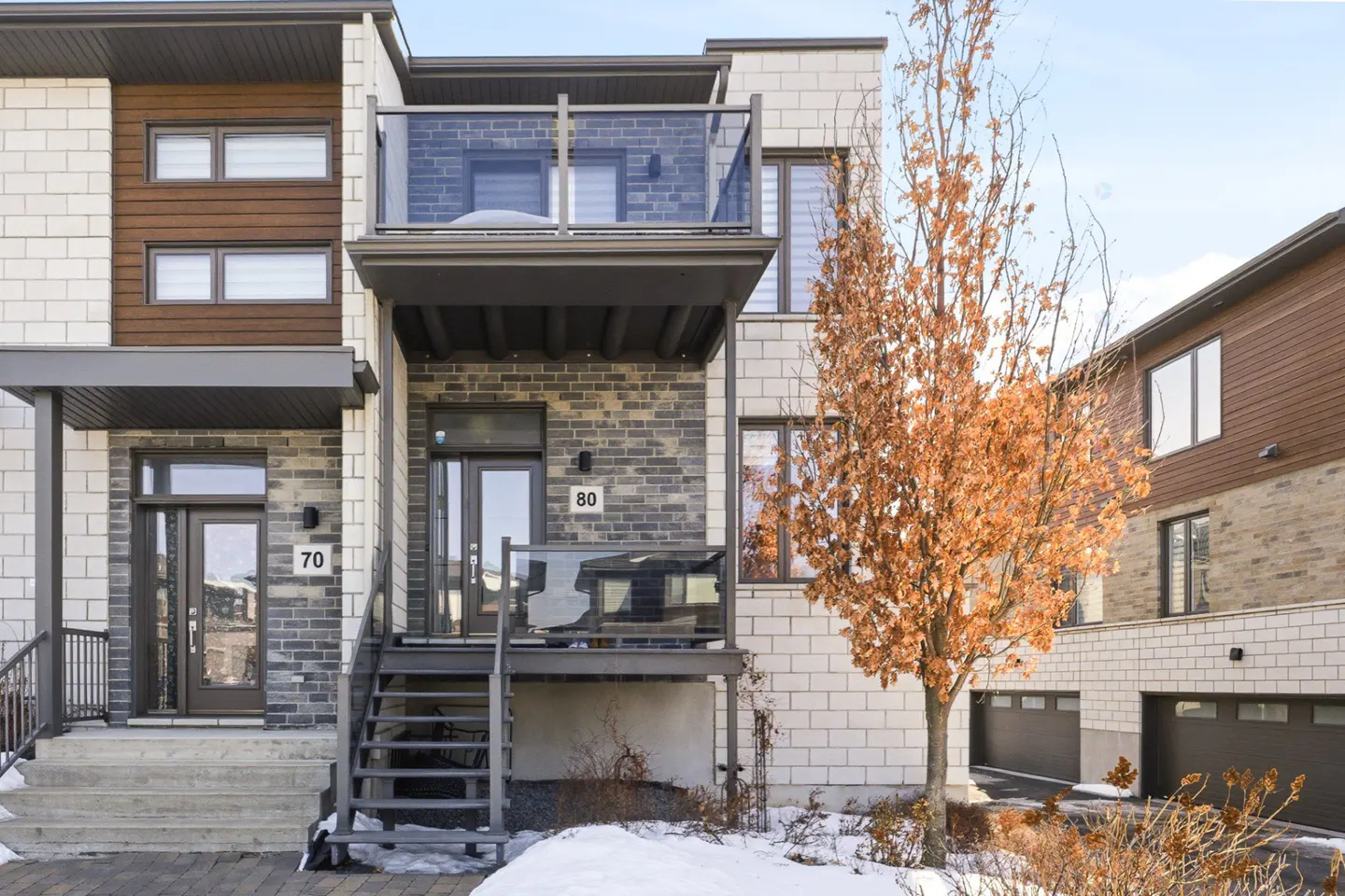 Exterior view of modern townhouses with brick and wood accents, featuring a tree with orange leaves and patches of snow.