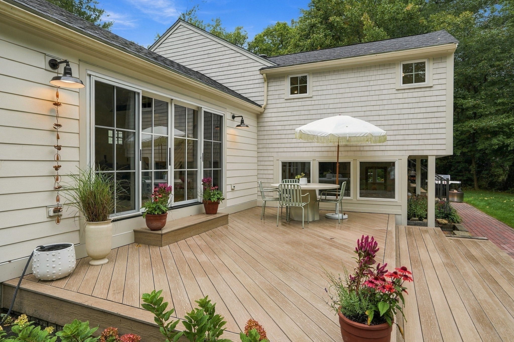 Spacious wooden deck with a dining set, white umbrella, and potted plants against a light-colored shingled house exterior. [AI-generated text]