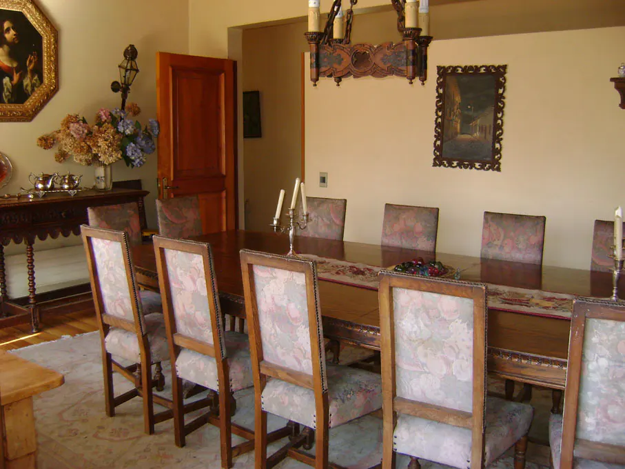 A dining room with a long wooden table and chairs with floral patterned upholstery. A chandelier hangs above the table.