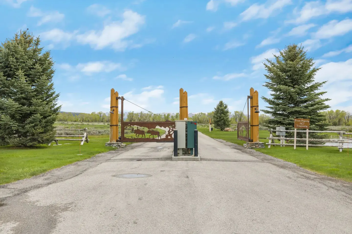 A paved driveway leads to a gated entrance with metal moose silhouettes, flanked by pine trees under a blue sky.