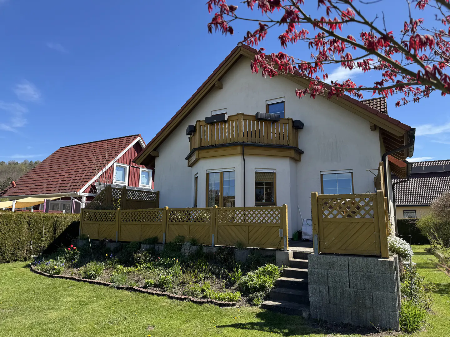 Two-story house with a balcony and a yellow fence, surrounded by a green lawn and blooming flowers. Red leaves on a tree are in the foreground.