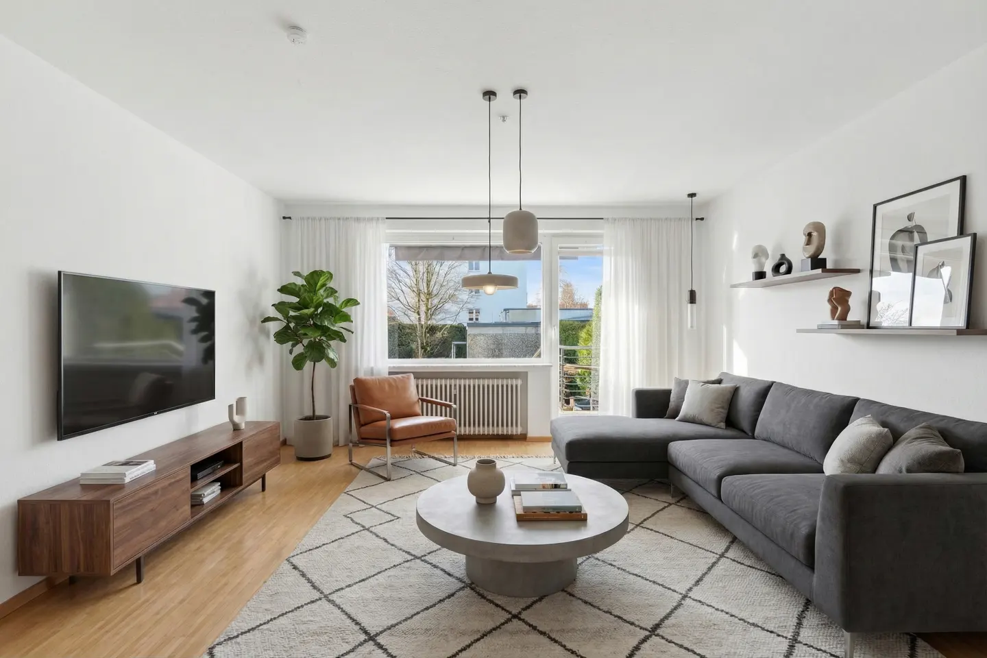 Bright living room with a gray sectional sofa, round coffee table, wood TV stand, and a leather chair near a window.