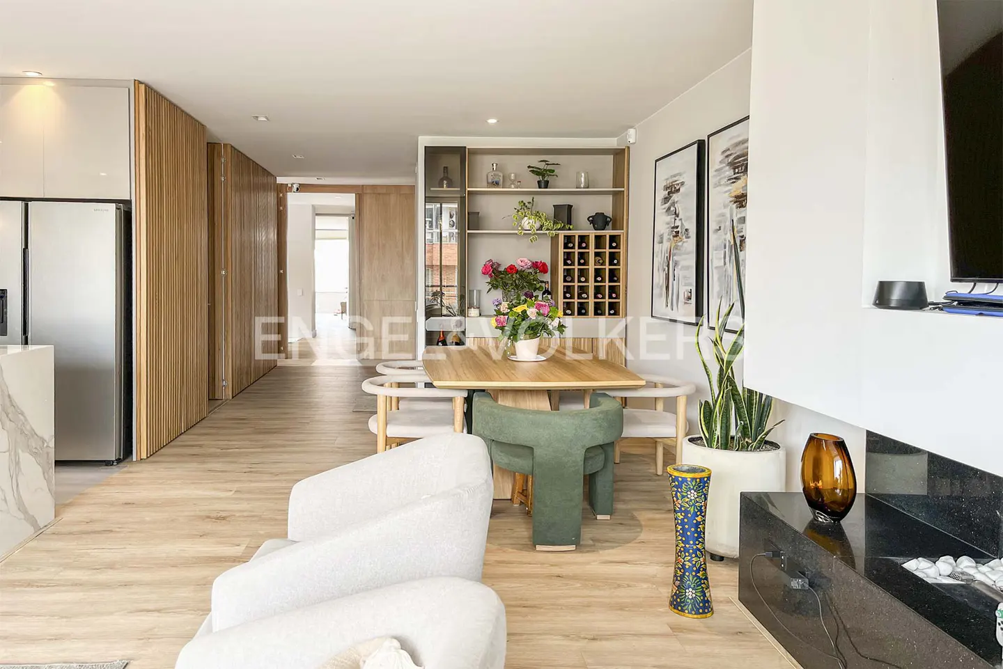 Bright, modern living and dining area with light wood floors, white sofa, and a wooden table with green chairs. A wine rack and art add detail.