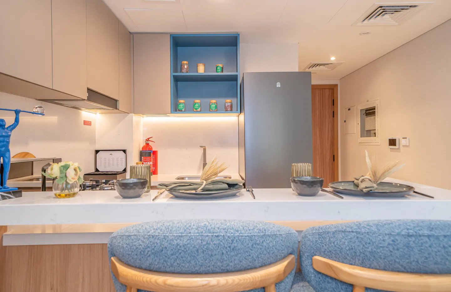 A modern kitchen with a white countertop, blue stools, and gray cabinets. A blue shelf displays jars. Place settings are on the counter.