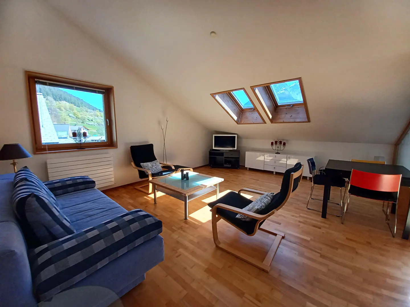 Attic living room with wood floors, blue sofa, chairs, table, TV, and dining set. Windows show mountain views.