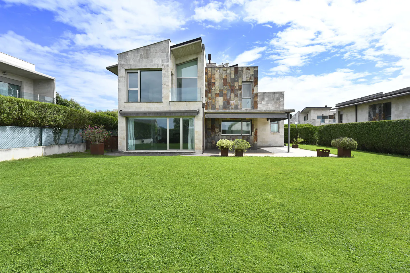 Two-story modern house with stone and glass exterior, surrounded by a lush green lawn under a blue sky with clouds.