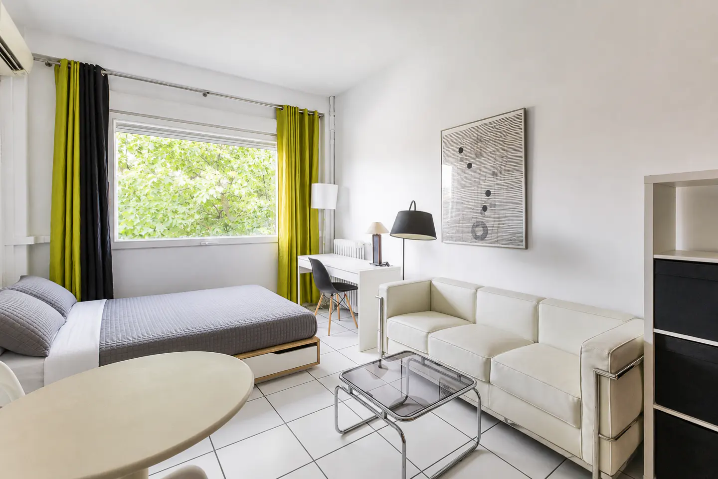 Bright studio apartment with white walls, tile floor, and modern furniture. A gray bed sits near a window with green curtains, and a white sofa faces a glass coffee table.