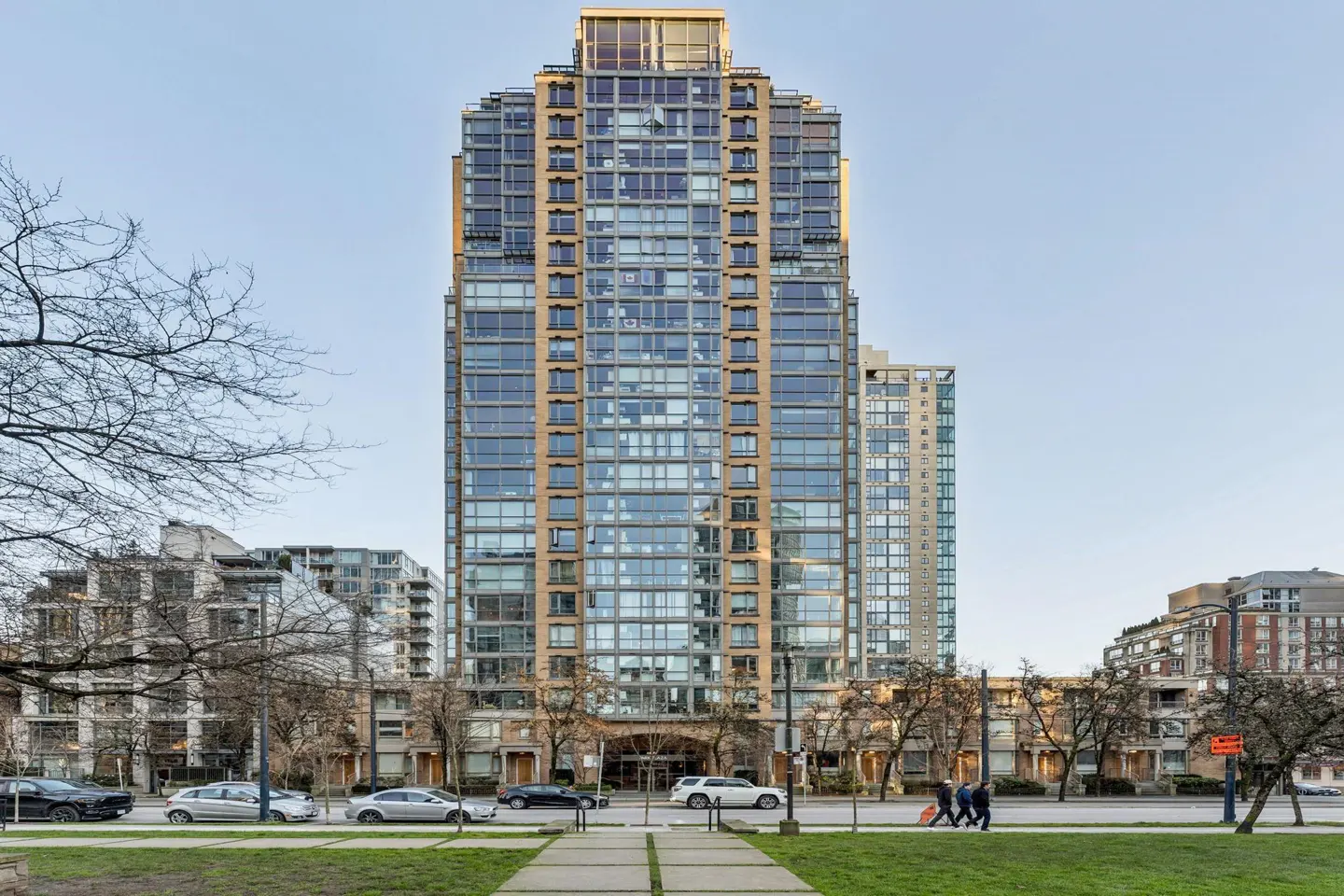 Exterior view of a tall, modern glass and tan-colored condo building with cars and people on the street.