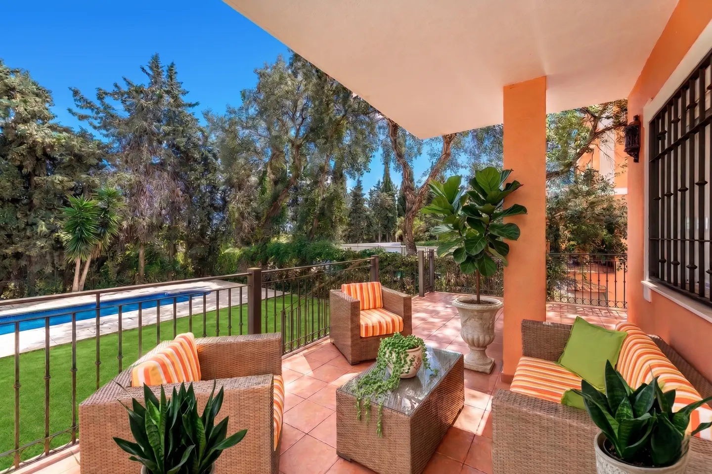 Outdoor patio with wicker furniture, orange striped cushions, and potted plants. A pool and trees are visible in the background.