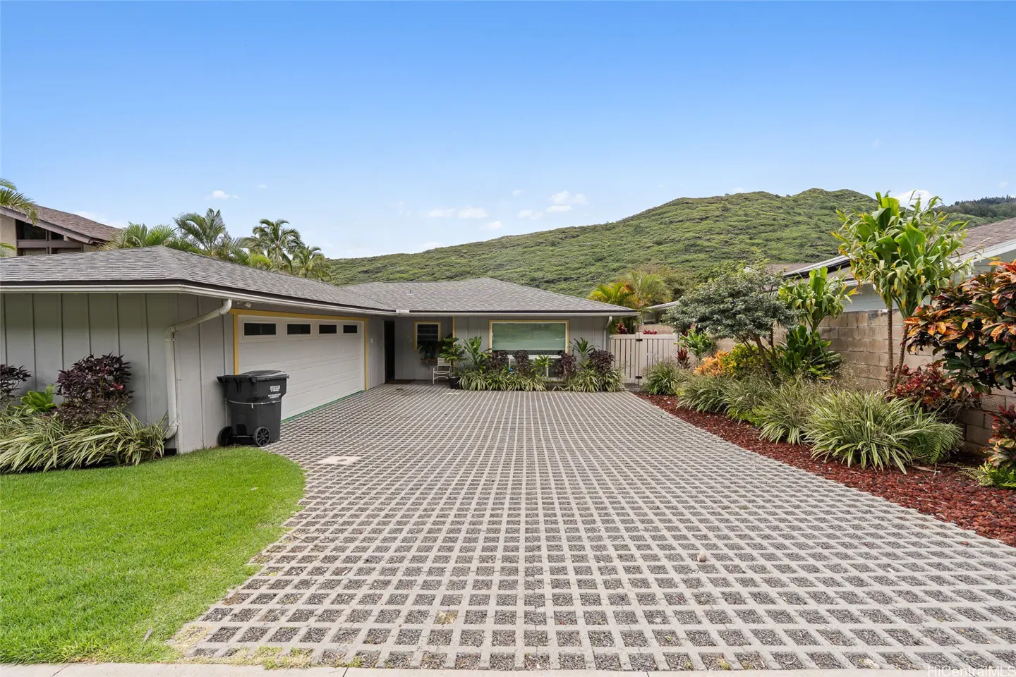 A single-story gray house with a white garage door and a patterned driveway under a blue sky. A green hill is in the background.