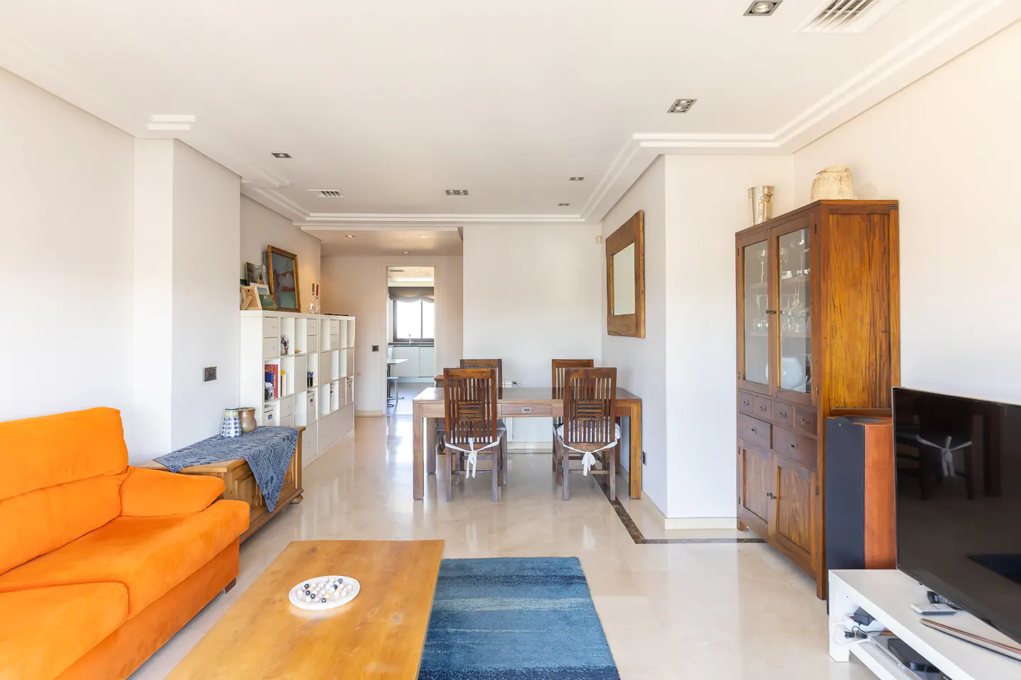 Bright living room with an orange sofa, wooden coffee table, blue rug, and a dining area with wooden chairs and a hutch.